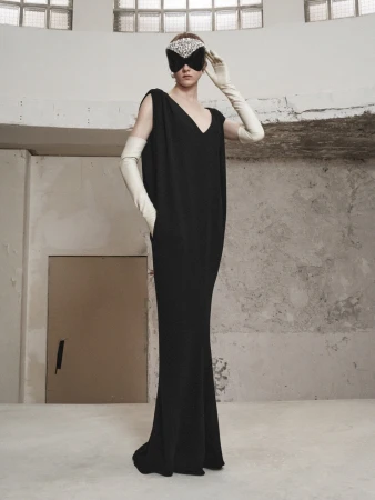 Fashion shot of a woman in a black dress and decorative mask, wearing long gloves, posing in a minimalist, industrial interior space.