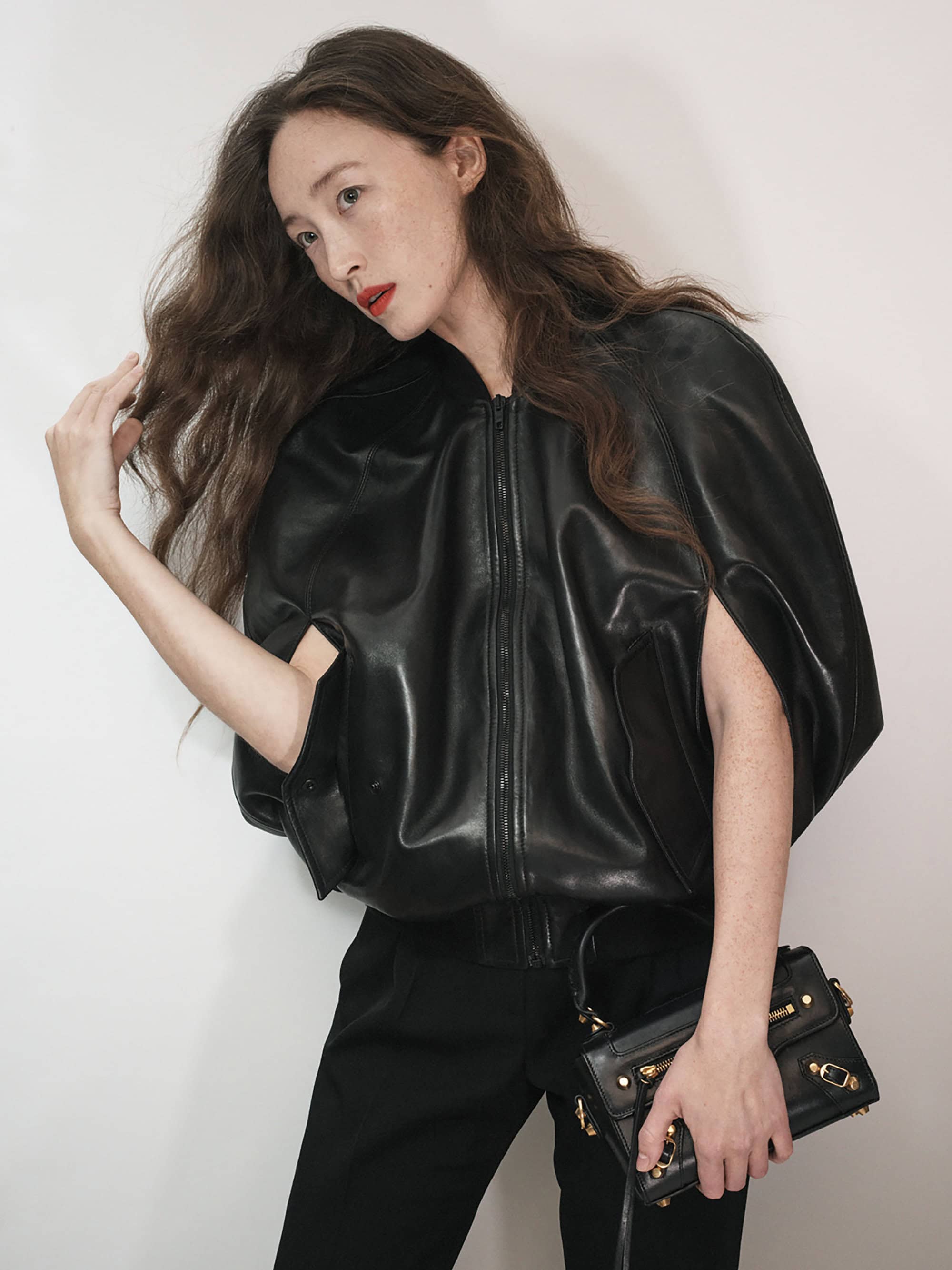 Adult female in black leather jacket and pants posing with a small studded leather handbag against a white background.