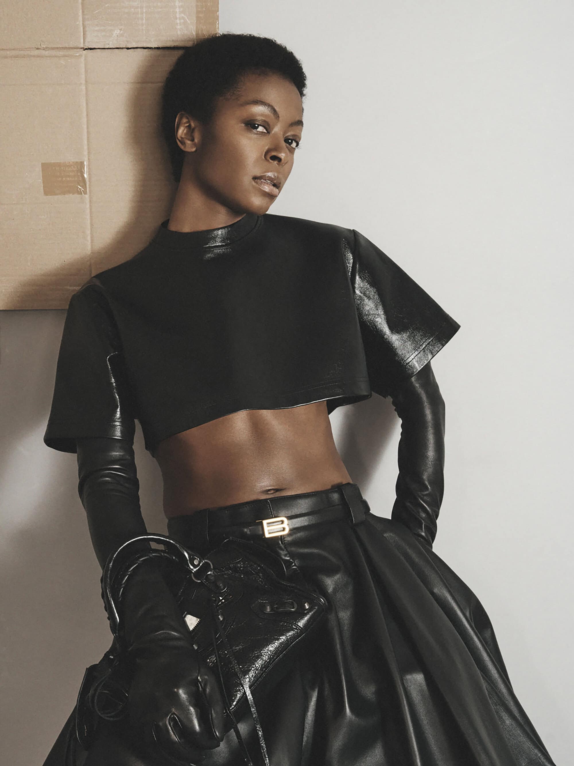 Fashion shot of a Black woman in a short black leather top and black leather skirt, posing indoors against a grey wall and brown cardboard.