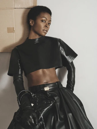 Fashion shot of a Black woman in a short black leather top and black leather skirt, posing indoors against a grey wall and brown cardboard.