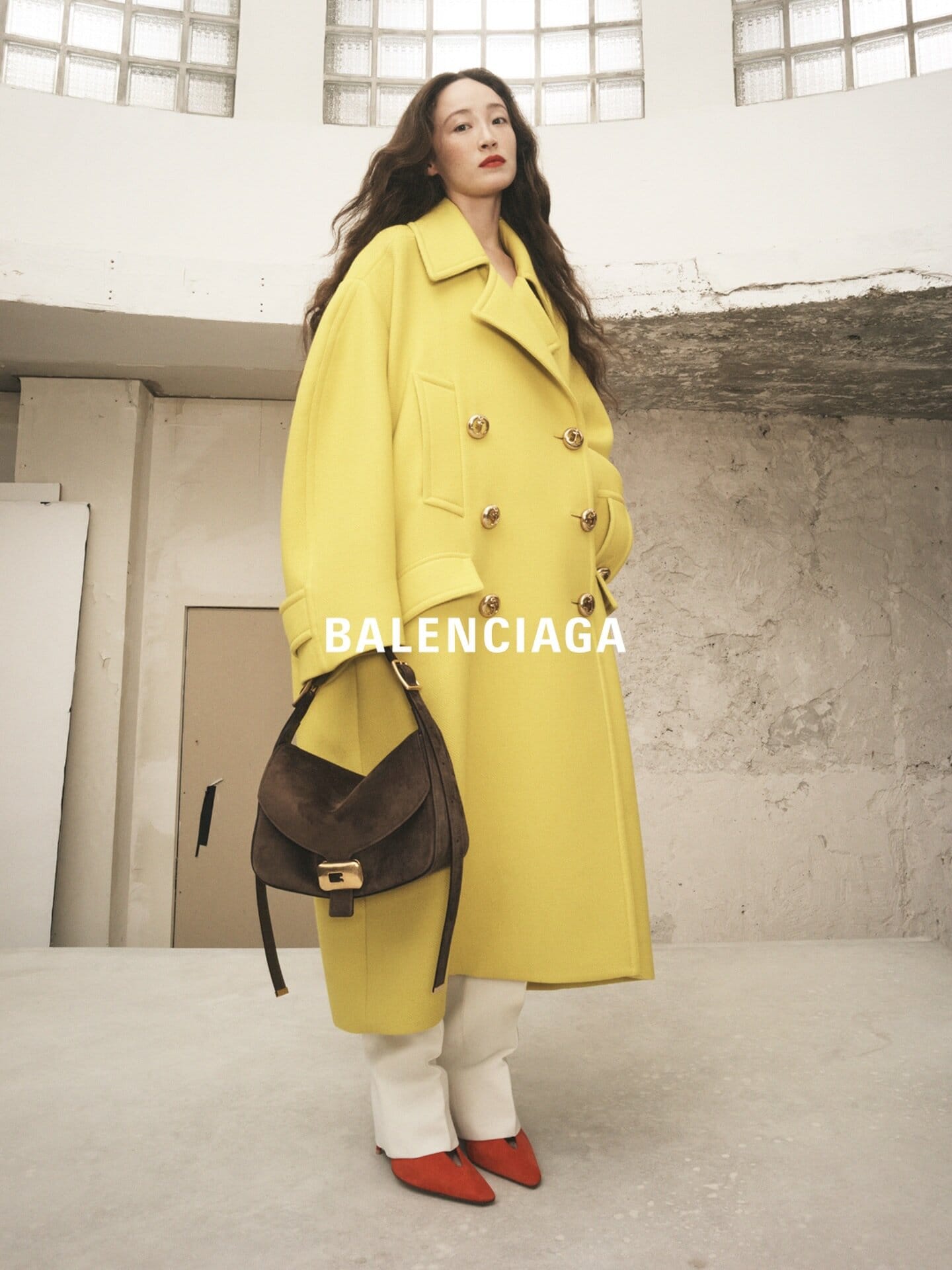 Elegant woman in a yellow Balenciaga coat holds a purse in a studio. She is wearing orange heels. Fashion editorial.