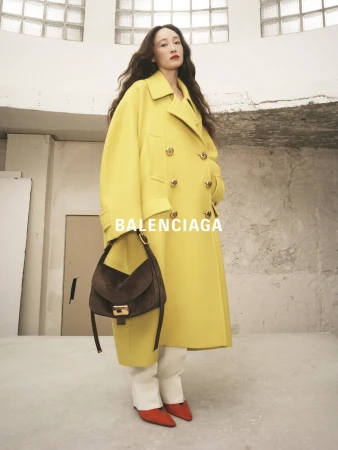 Elegant woman in a yellow Balenciaga coat holds a purse in a studio. She is wearing orange heels. Fashion editorial.
