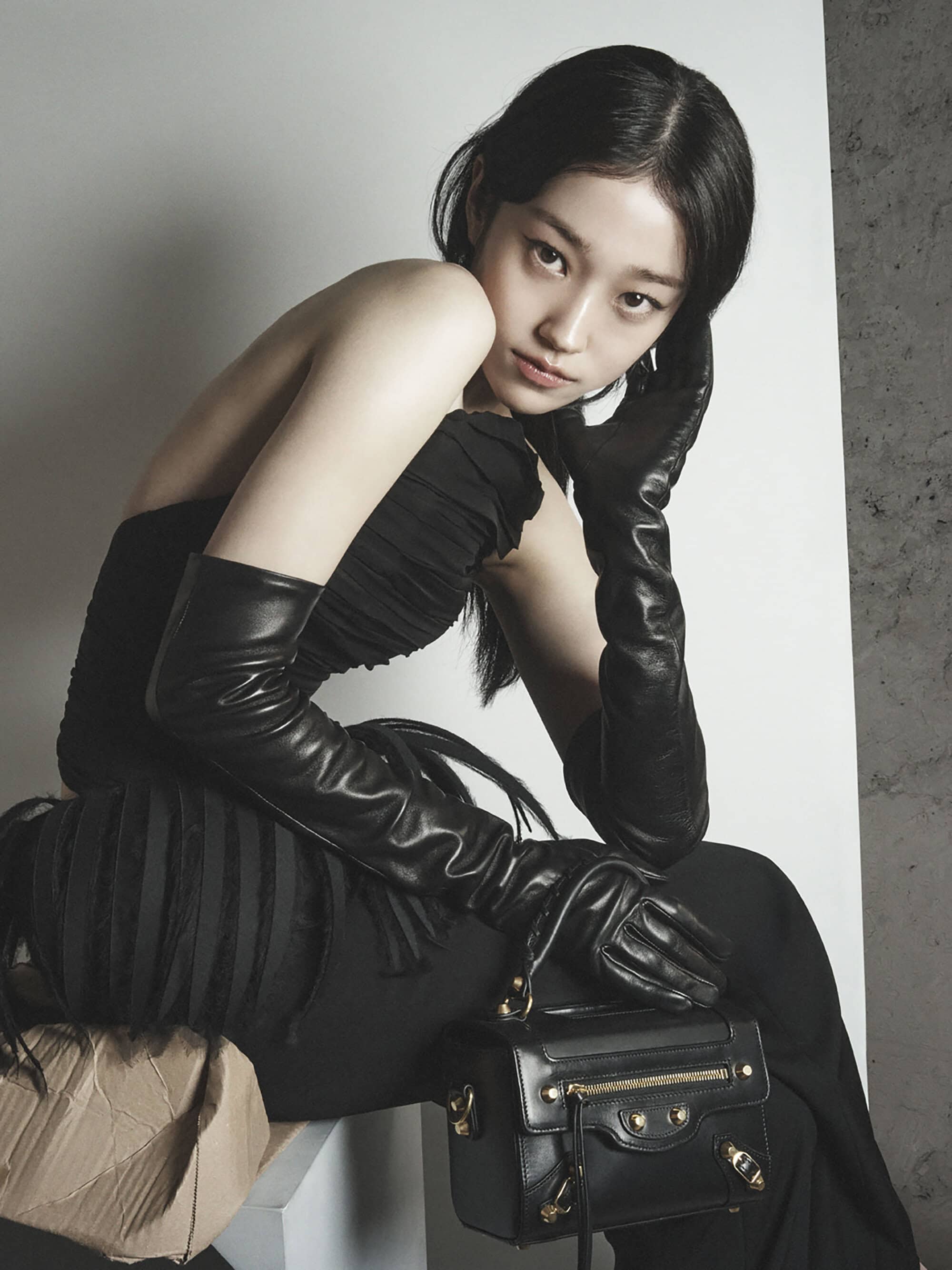 An Asian woman poses in a black strapless dress and long black gloves, sitting on a white cube with a small black purse beside her.