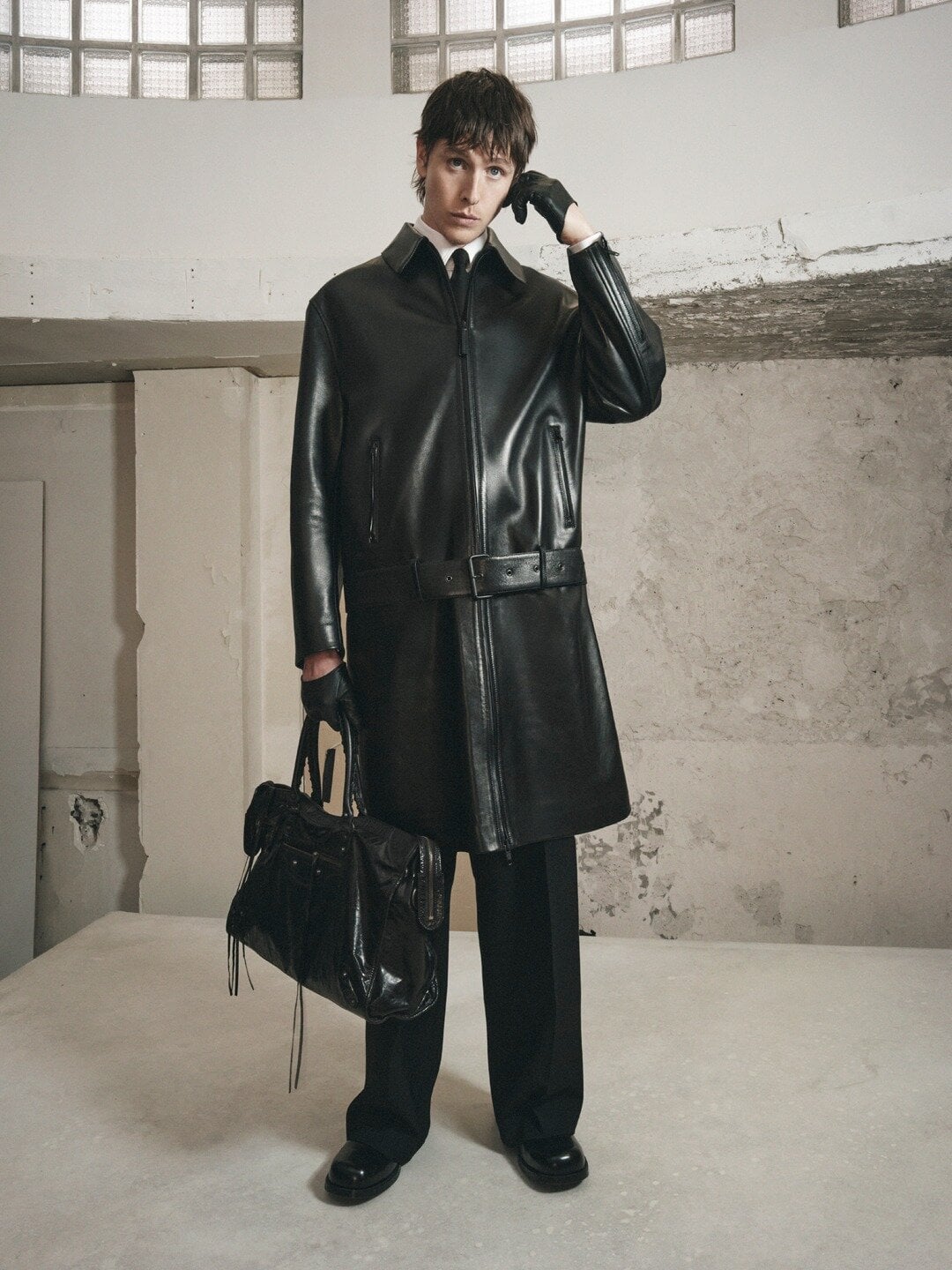 Adult male poses indoors wearing a long black leather jacket, gloves, black pants, and shiny shoes holding a large black leather bag.