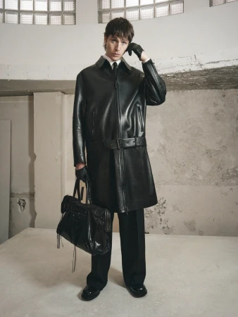 Adult male poses indoors wearing a long black leather jacket, gloves, black pants, and shiny shoes holding a large black leather bag.