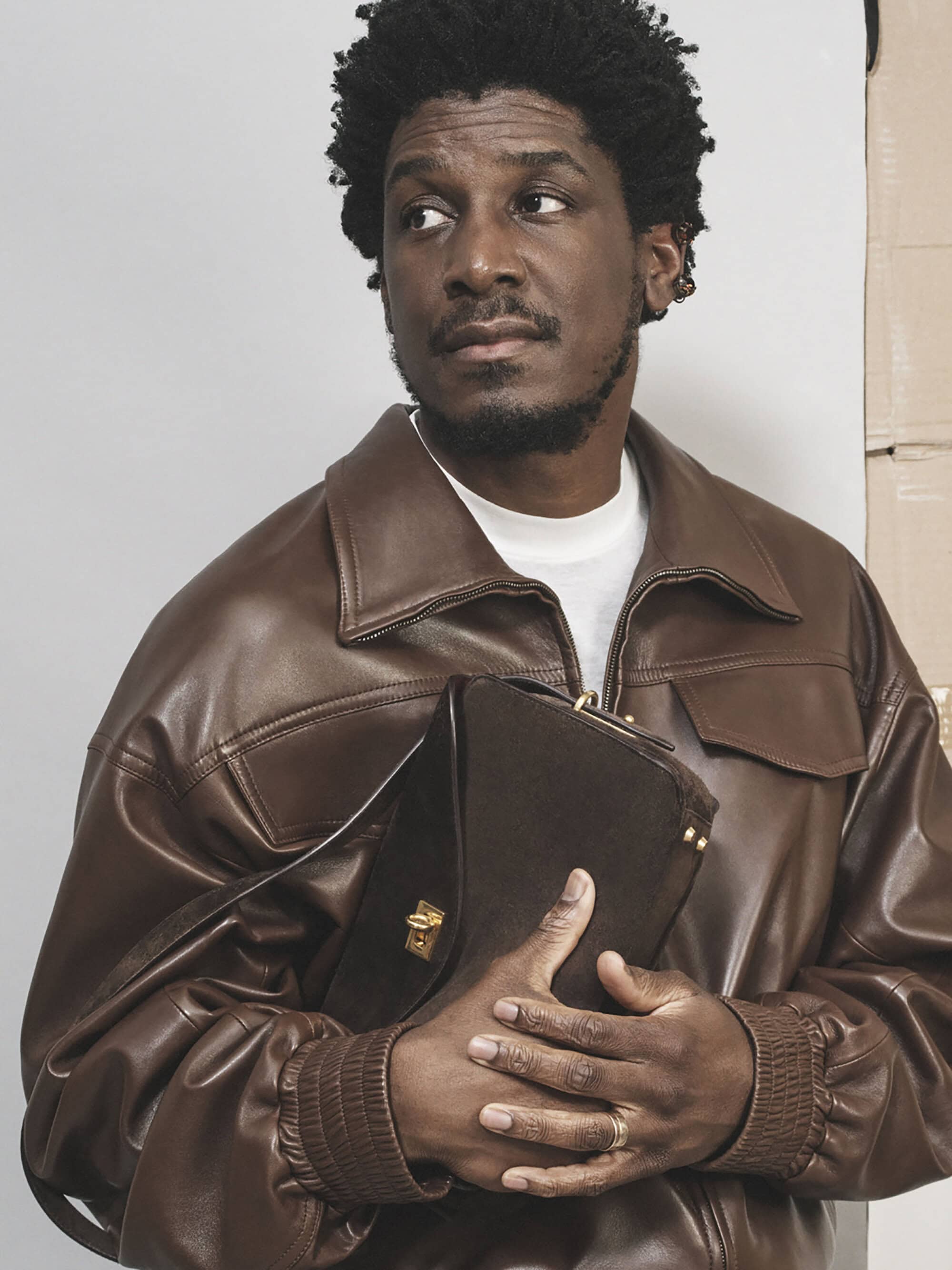 An adult Black man with curly hair in a brown leather jacket, holding a brown purse, looking left, in a studio setting with a neutral backdrop.