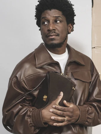 An adult Black man with curly hair in a brown leather jacket, holding a brown purse, looking left, in a studio setting with a neutral backdrop.