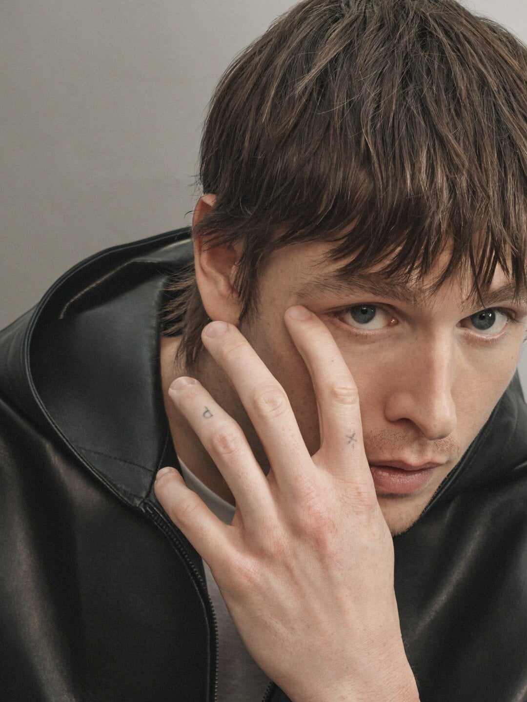 Close-up portrait of a young adult male with brown hair and blue eyes, wearing a black hooded jacket, with his hand near his face.