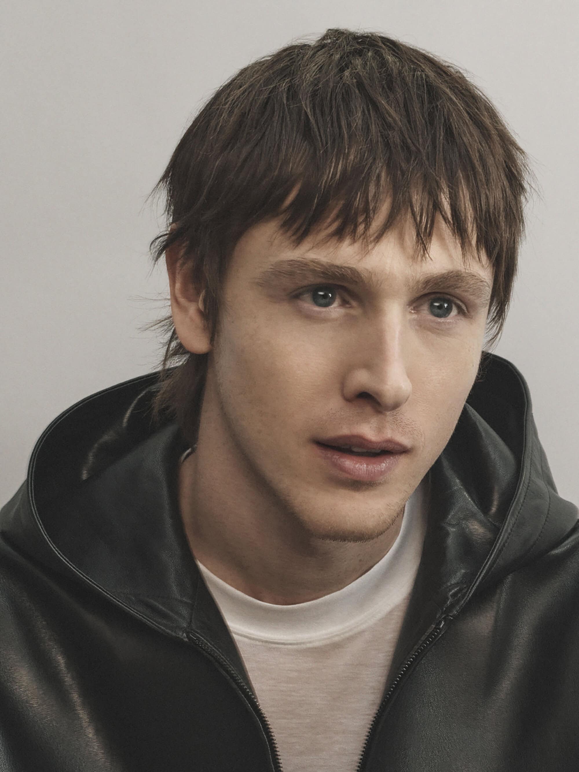 Portrait of a young adult male with brown hair wearing a black leather hooded jacket and a white t-shirt, posing against a neutral background.