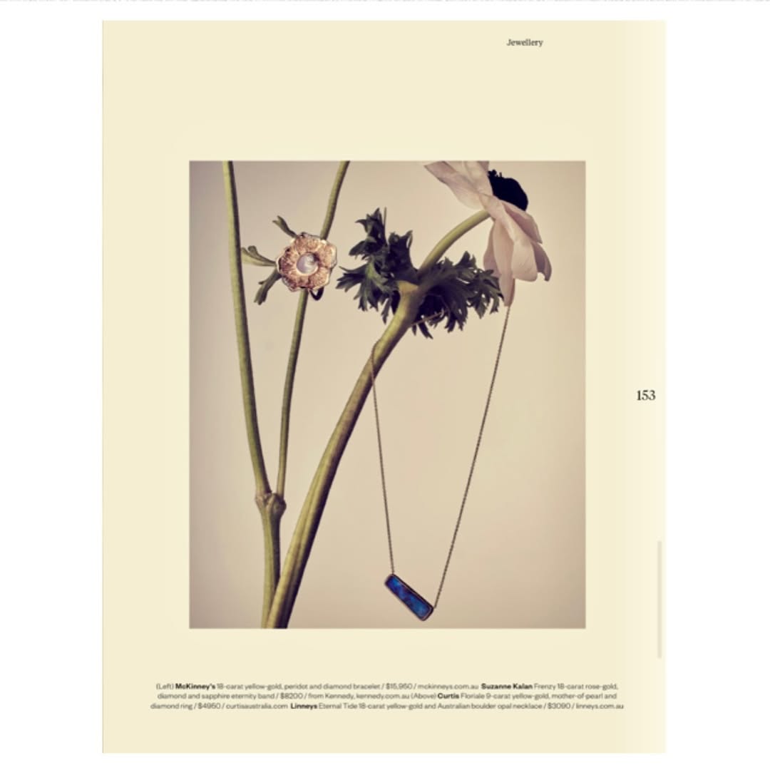 Editorial fashion shot of gold jewelry featuring a ring and necklace alongside a flowering plant against a beige background.