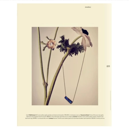 Editorial fashion shot of gold jewelry featuring a ring and necklace alongside a flowering plant against a beige background.