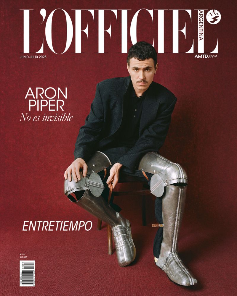 Aron Piper sits on a stool wearing a dark suit jacket and metallic leg armor against a red background for a magazine cover.