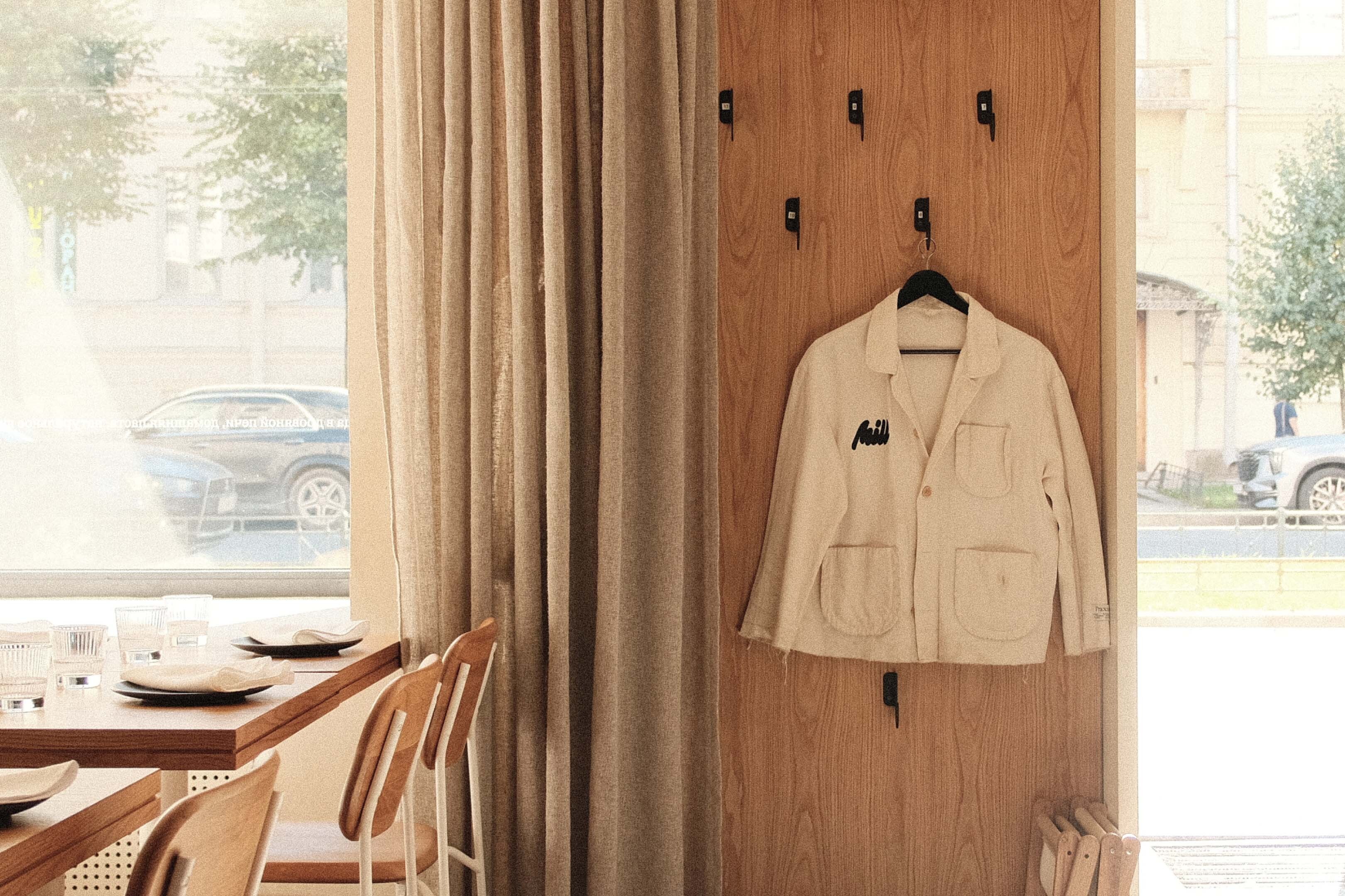 Beige jacket hangs on a wooden wall with metal hooks inside a restaurant, accompanied by tables, chairs, curtains, and a street view.