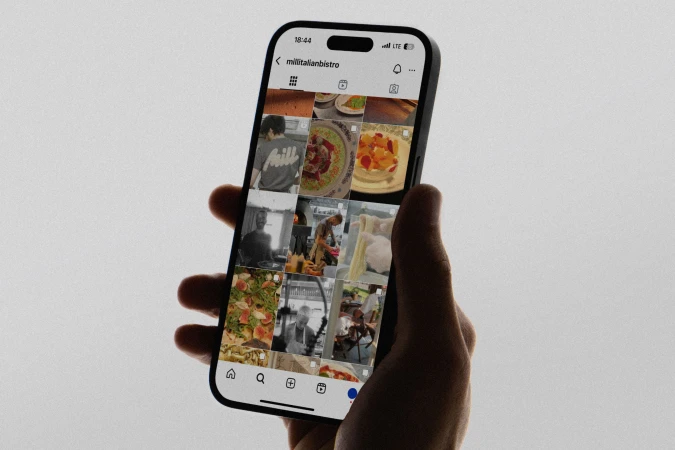 Hand holding a smartphone displaying an Instagram feed with food and people photos, set against a plain background in soft lighting.