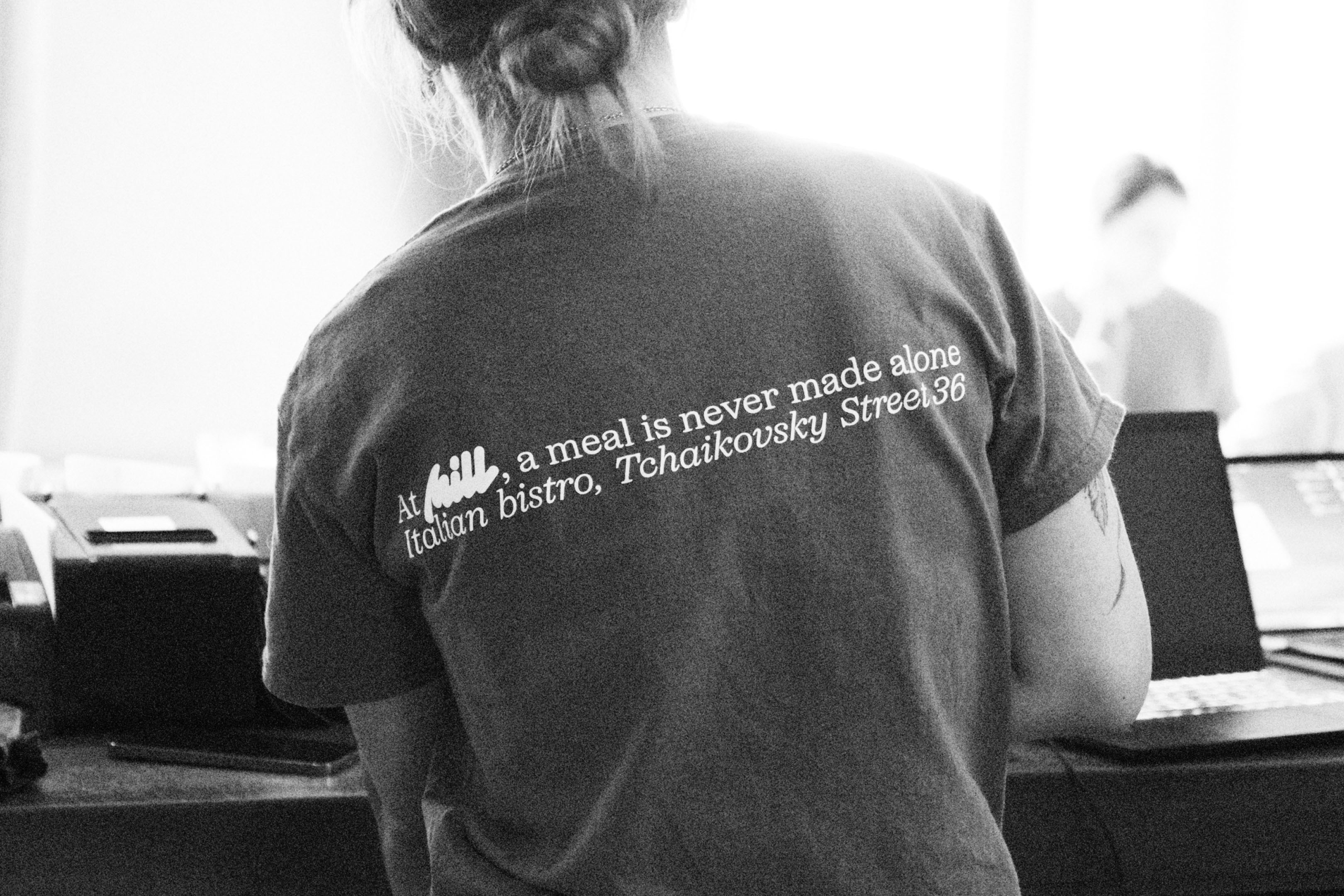 Back view of a person in a t-shirt with restaurant text standing indoors, with a blurred figure and objects in the background.
