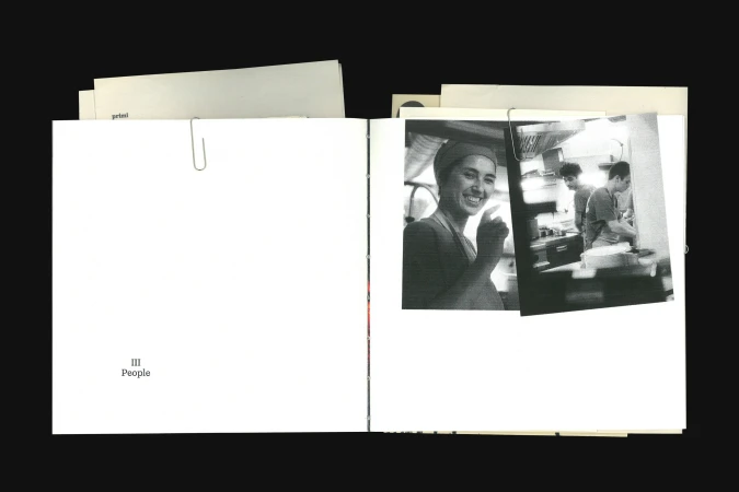 Monochrome image inside a book showcasing an adult woman smiling in a kitchen, with two adults working in the background.