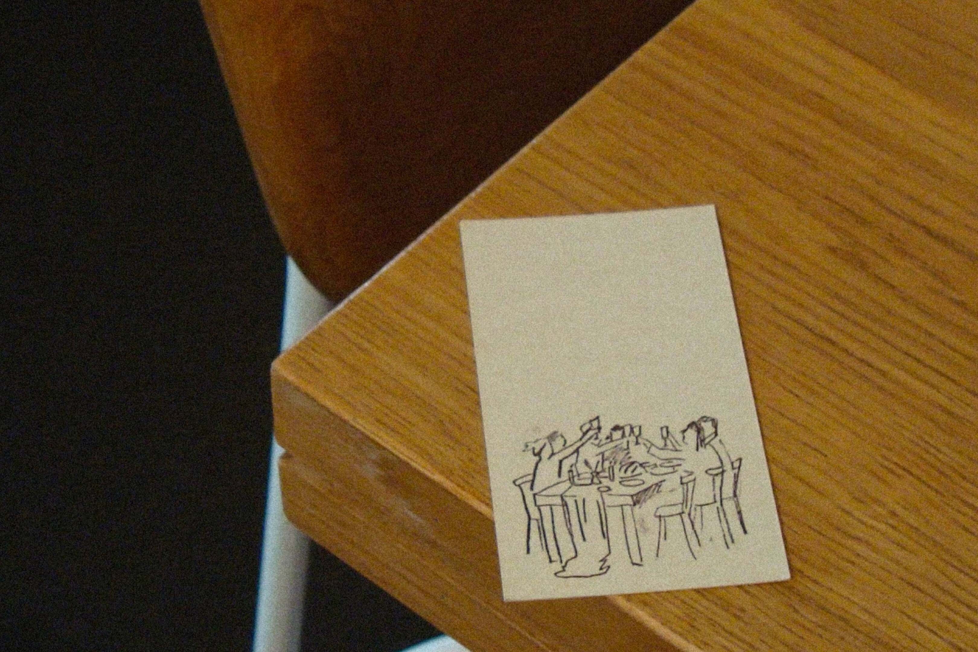 Close-up of a sketch on a card on a wooden table. The sketch depicts figures seated around a table, rendered in simple black lines.