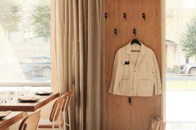 Off-white work jacket hanging on a wooden wall panel inside a contemporary interior setting with a table and chairs.