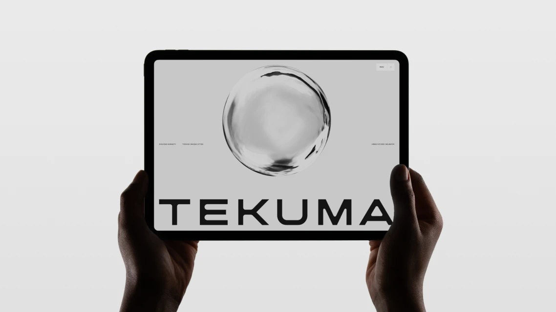 Hands hold a black tablet displaying a website with a grayscale abstract sphere and the name TEKUMA in bold letters on a light gray screen.