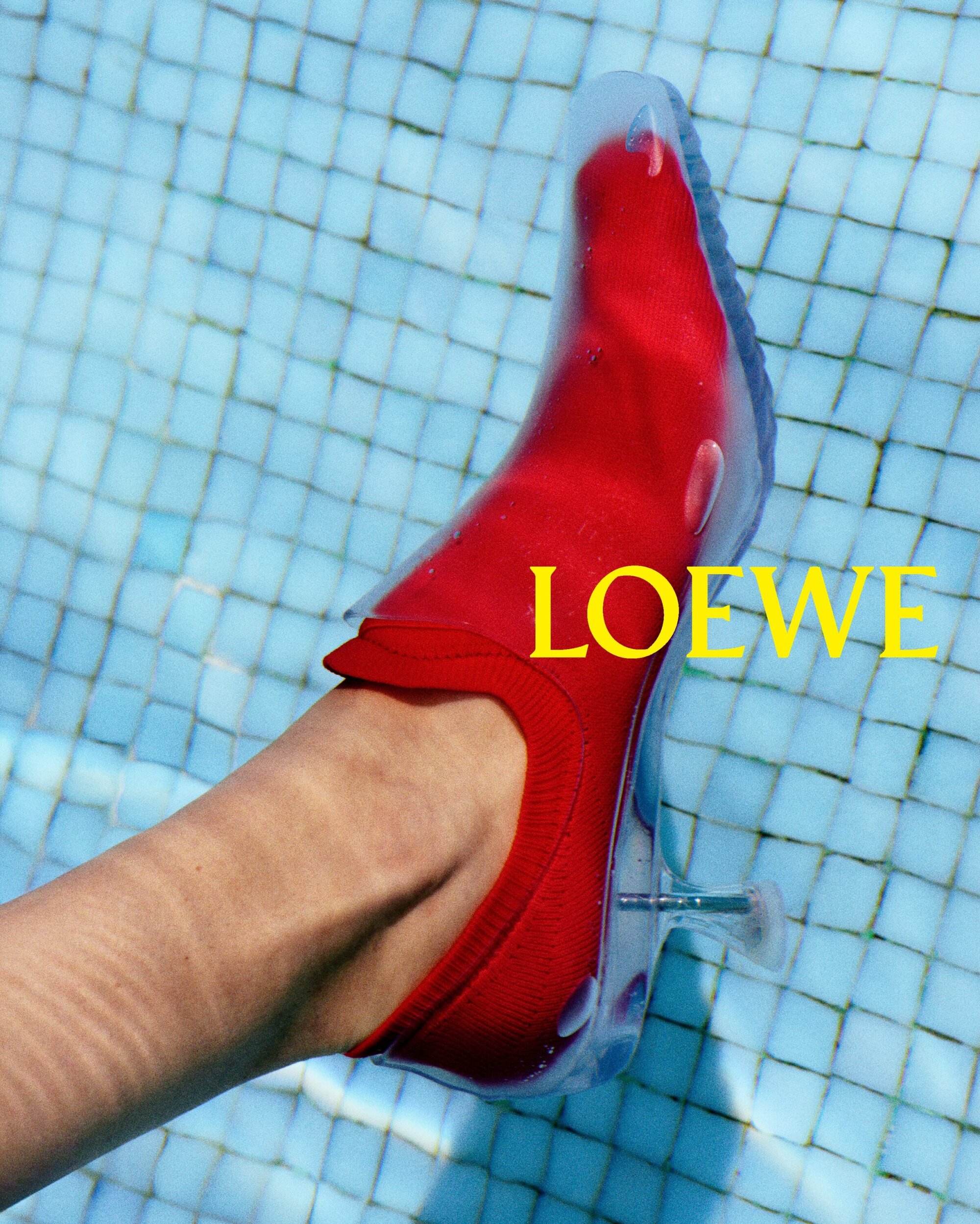 A person's foot and red high-heeled shoe are partially submerged in a pool with blue tiles, featuring the yellow 'LOEWE' logo.
