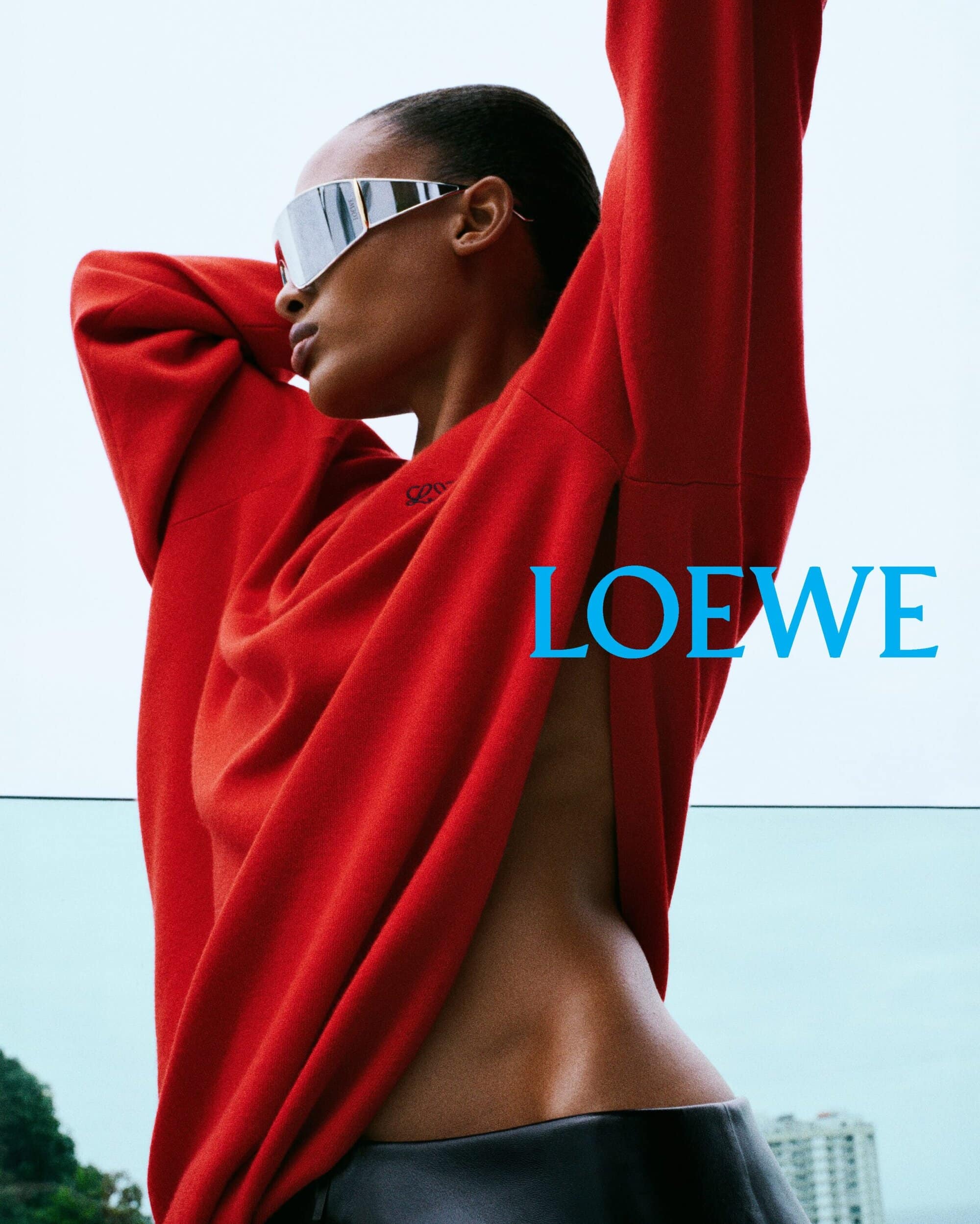 Model in reflective visor and red sweatshirt raising arms outdoors with LOEWE text visible