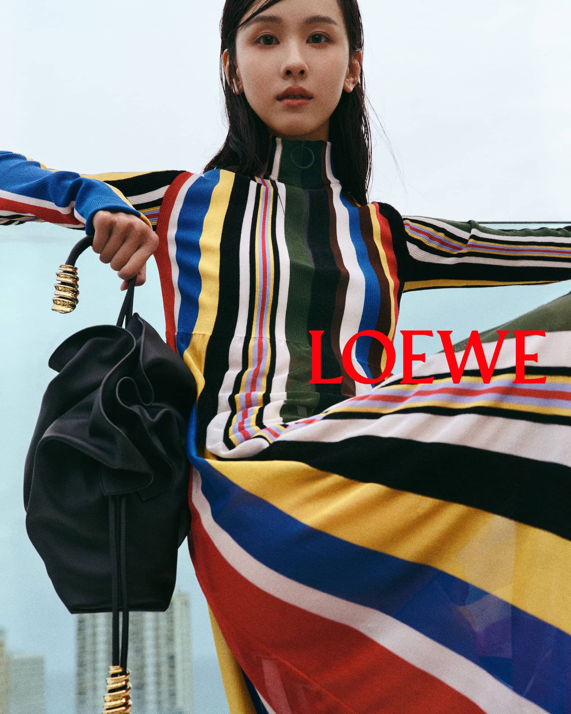 Elegant Asian woman poses outdoors in a striped dress, holding a black drawstring bag, against a blurred city skyline backdrop.