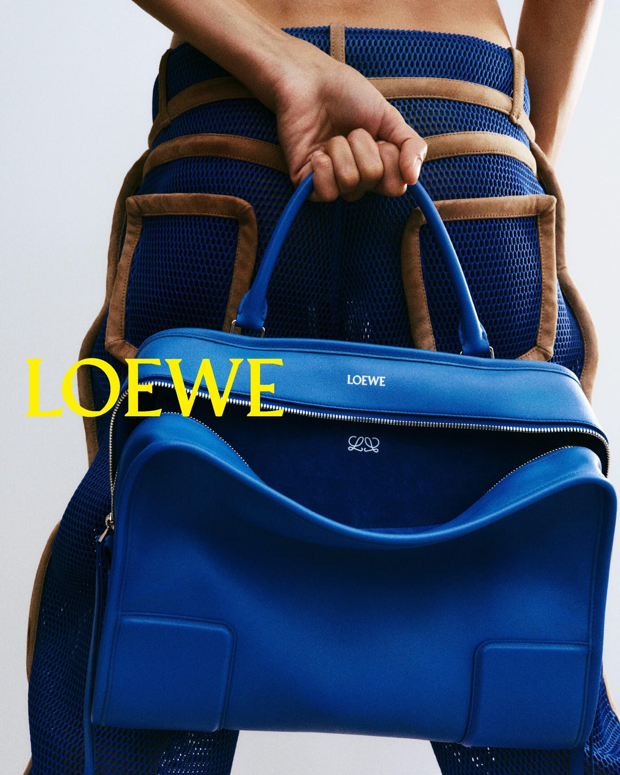 Close-up studio shot of a blue leather Loewe handbag held by a slender woman with bare midriff wearing matching blue apparel.