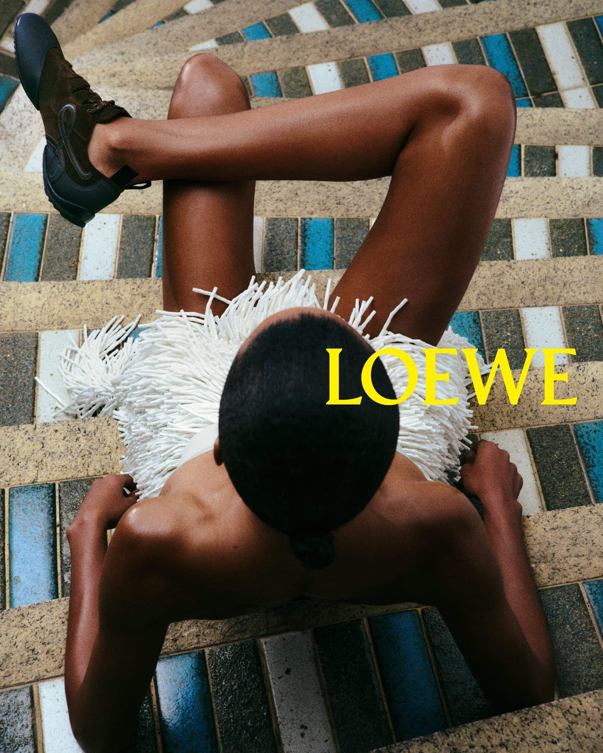 An adult with dark skin poses on a staircase wearing brown shoes and a white fringe garment, the brand name LOEWE is overlaid in yellow.