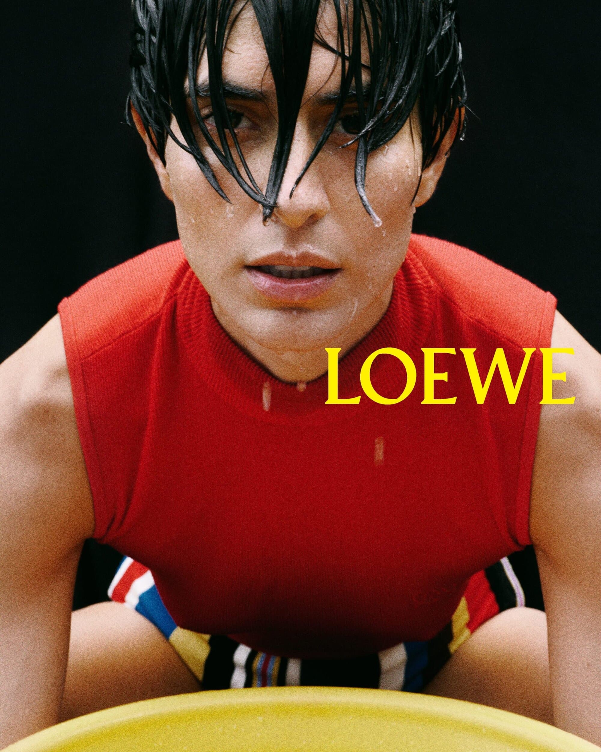 Close-up of a man in a red sleeveless shirt with wet hair, posing indoors with the 'LOEWE' logo visible in yellow text.
