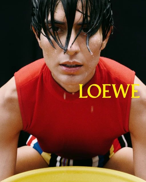 Close-up of a man in a red sleeveless shirt with wet hair, posing indoors with the 'LOEWE' logo visible in yellow text.
