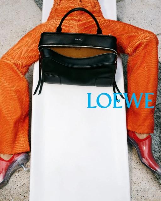 Fashionable handbag presented on a white surface, paired with orange textured pants and transparent heeled shoes, with Loewe logo.