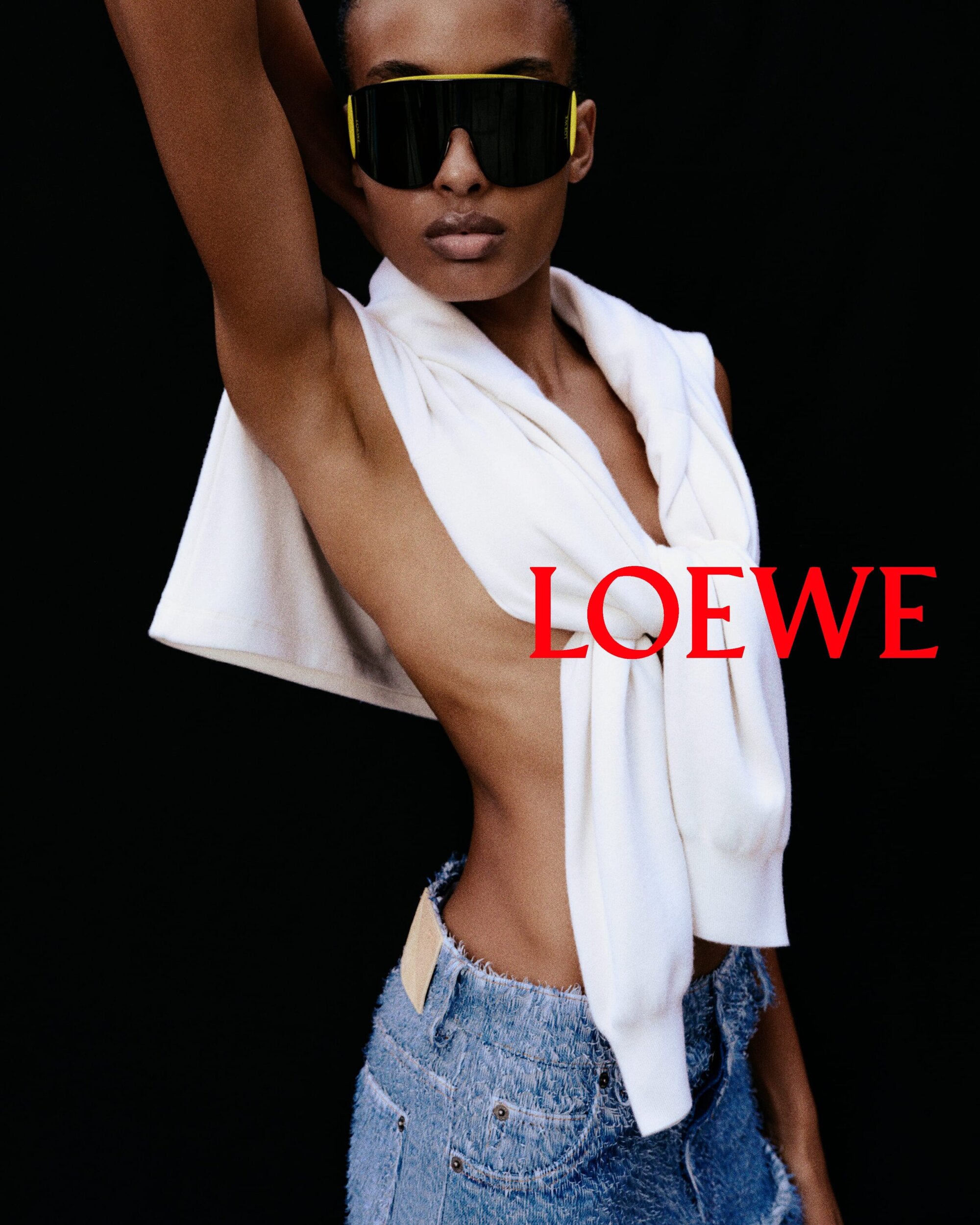 Model wearing oversized black sunglasses and a white sweater tied around shoulders with denim shorts against black backdrop