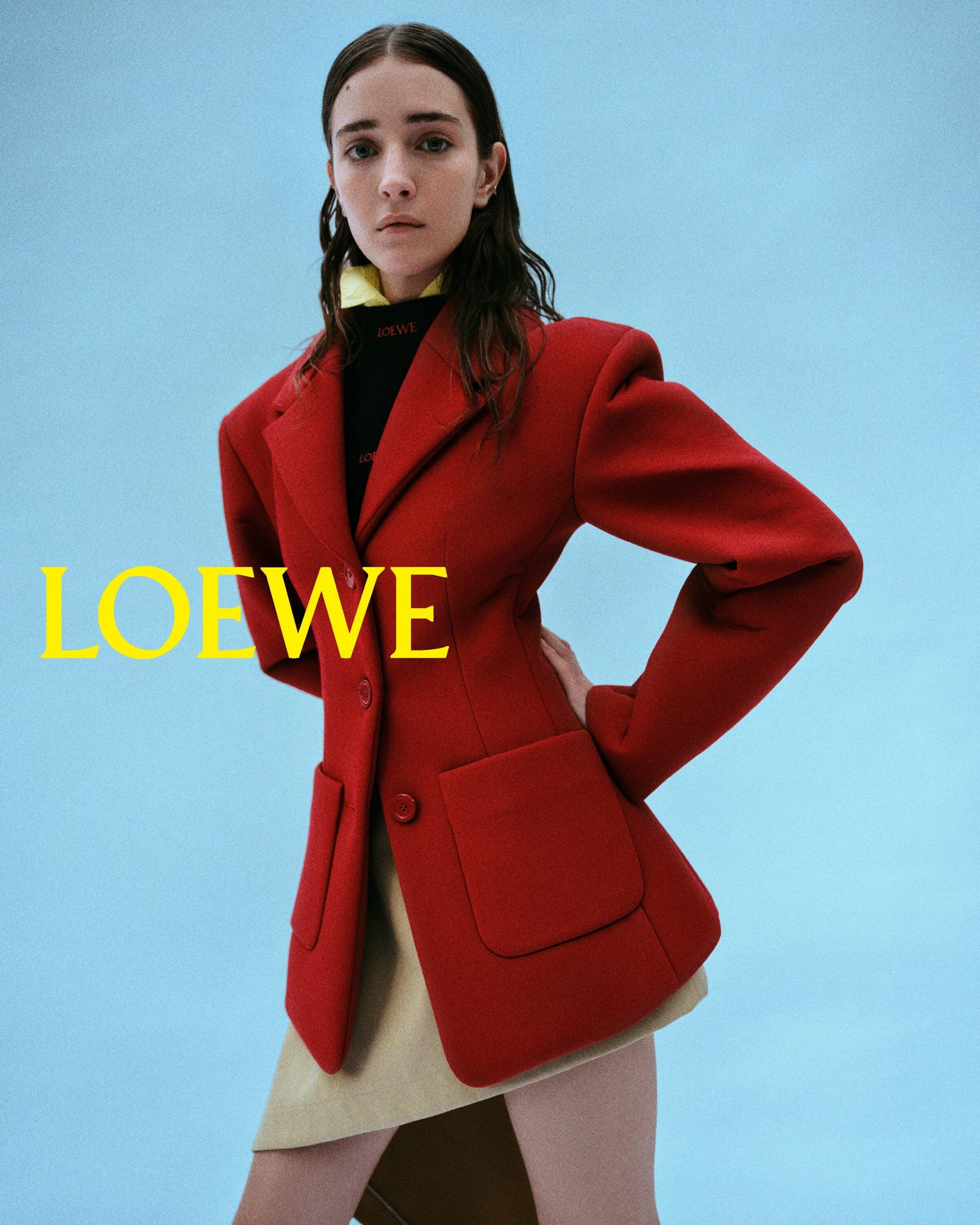 Fashion editorial shot of a woman in a red blazer and tan skirt against a light blue background, with the 'Loewe' logo in view.