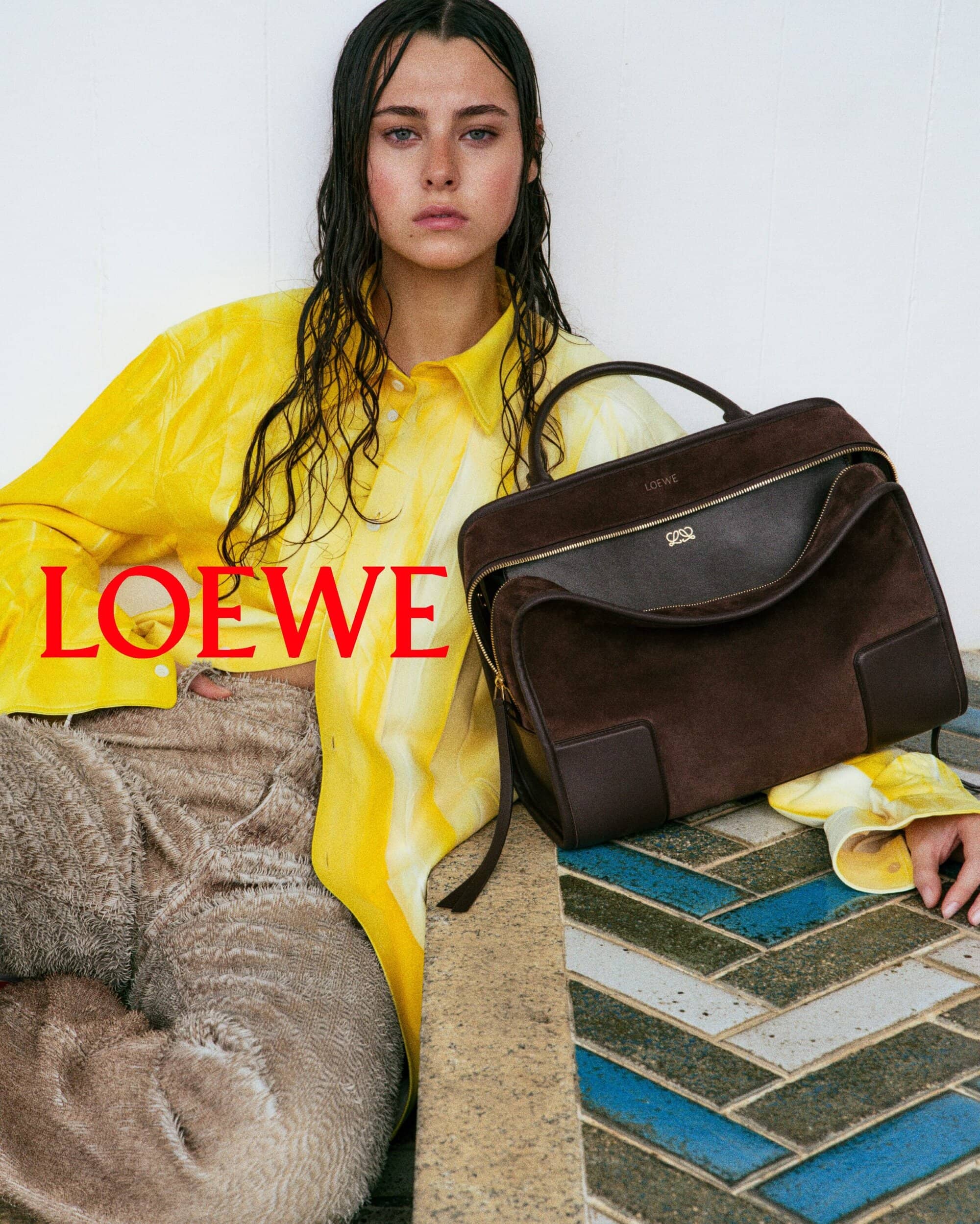 Fashion shot of a woman with a brown handbag and a yellow tie-dye shirt in a studio with a patterned tile floor. Loewe logo is visible.
