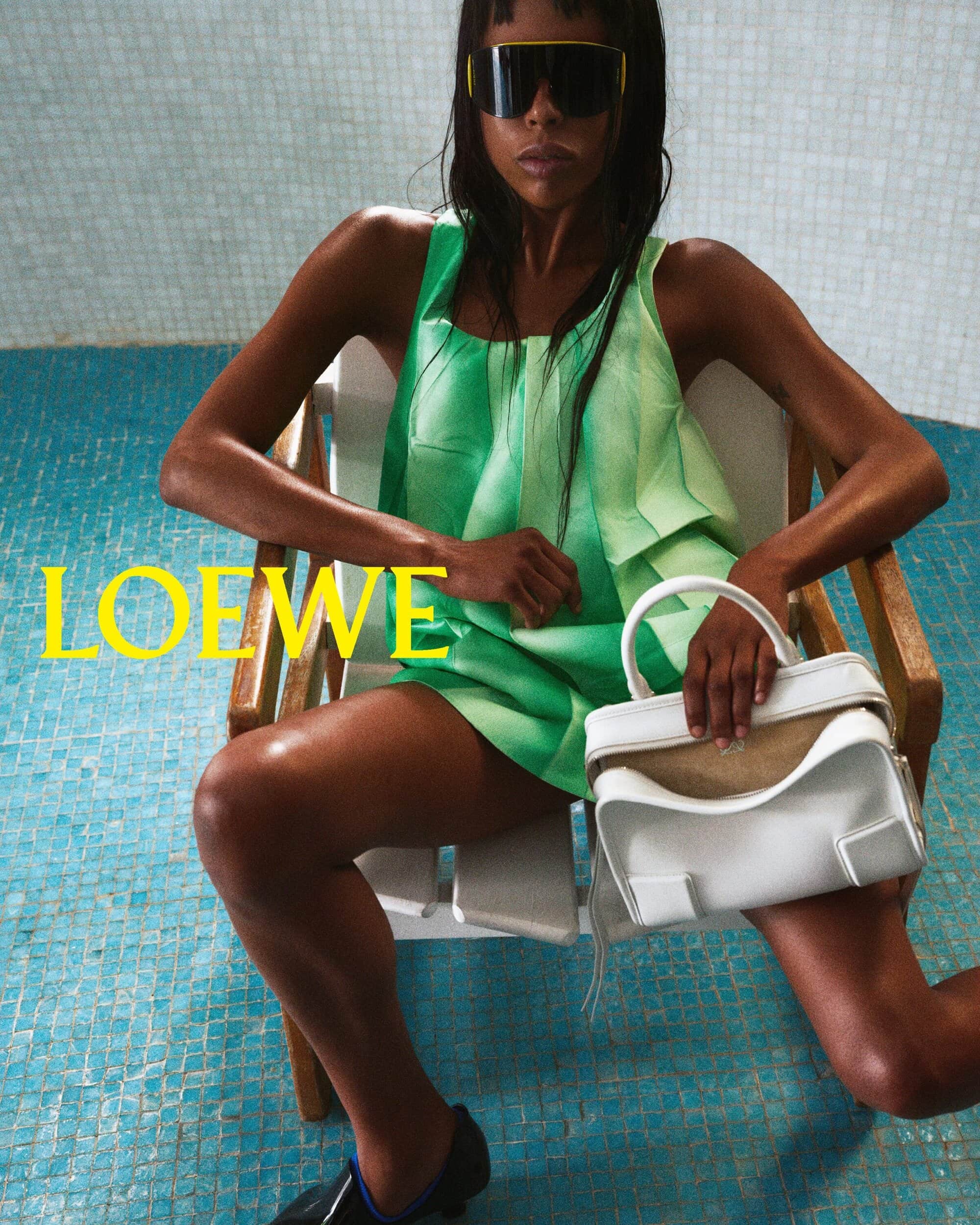 Fashion shot of a Black woman in green outfit, sunglasses, holding a white purse, seated on a white lounge chair with blue tiled pool background.