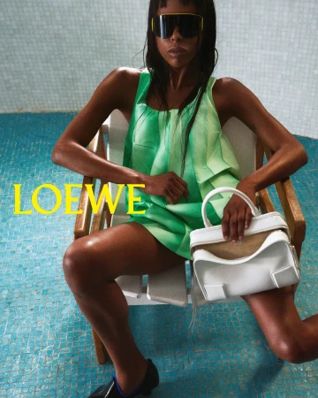 Fashion shot of a Black woman in green outfit, sunglasses, holding a white purse, seated on a white lounge chair with blue tiled pool background.