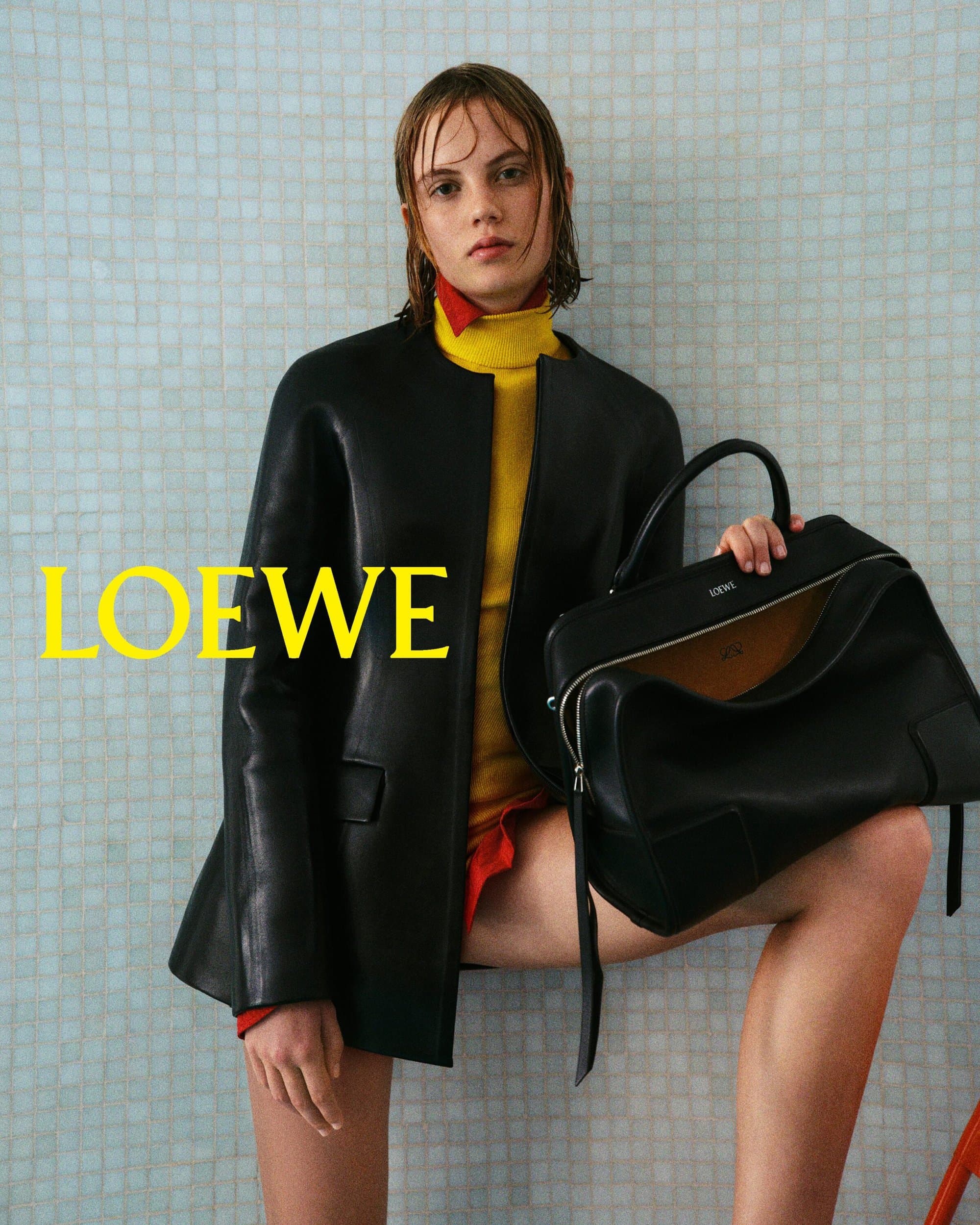 A woman poses in a black jacket with a large black leather bag in a studio with a minimalist tiled backdrop for a fashion shoot.