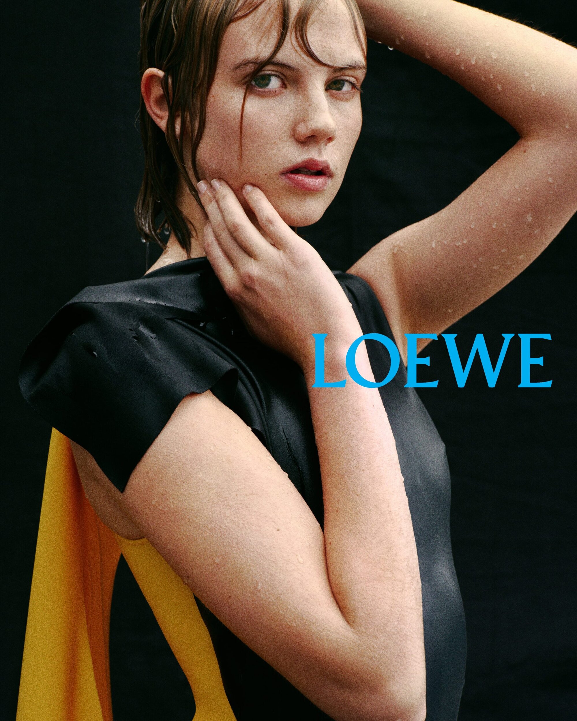 Close-up fashion portrait of a young woman with wet hair, wearing a black top and yellow drape, posing against a dark background.