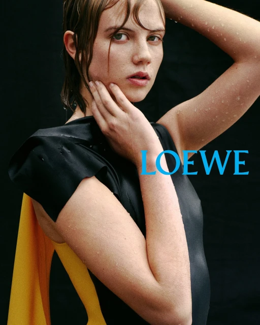 Close-up fashion portrait of a young woman with wet hair, wearing a black top and yellow drape, posing against a dark background.