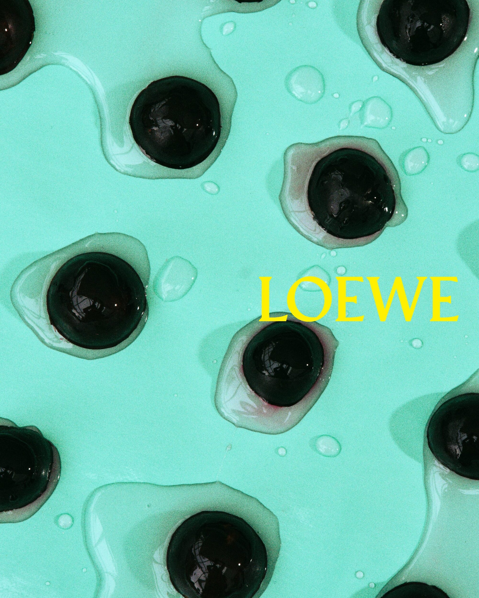 Overhead shot of black cherries covered with a shiny glaze on a light blue background with the text overlay “LOEWE” in yellow.
