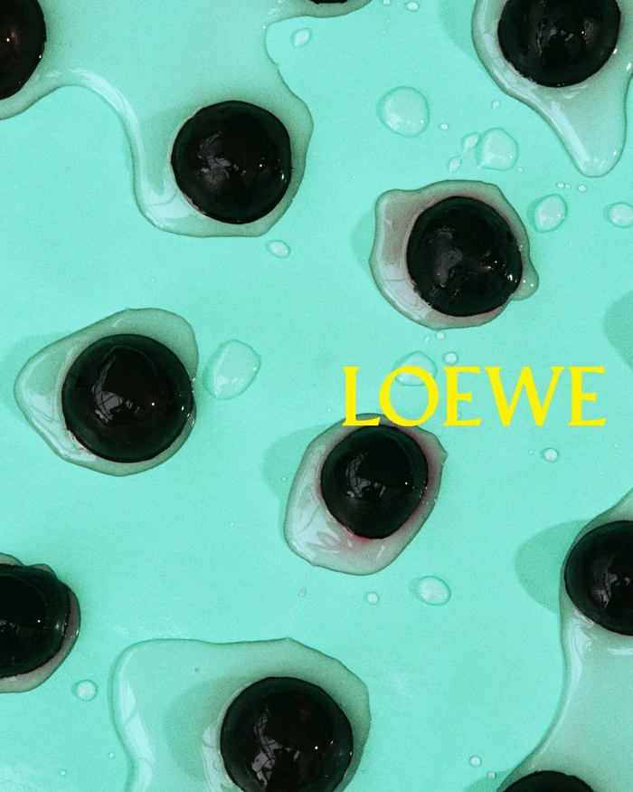 Close-up shot of dark spherical objects resting in puddles of clear liquid on a vibrant turquoise background with the Loewe brand name.