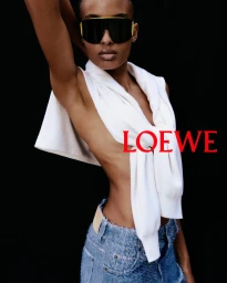 Model wearing oversized black sunglasses and a white sweater tied around shoulders with denim shorts against black backdrop