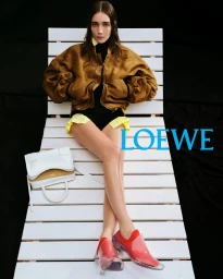 Model seated on a white wooden lounger wearing a brown bomber jacket, black bodysuit, and red shoes with a white handbag beside her