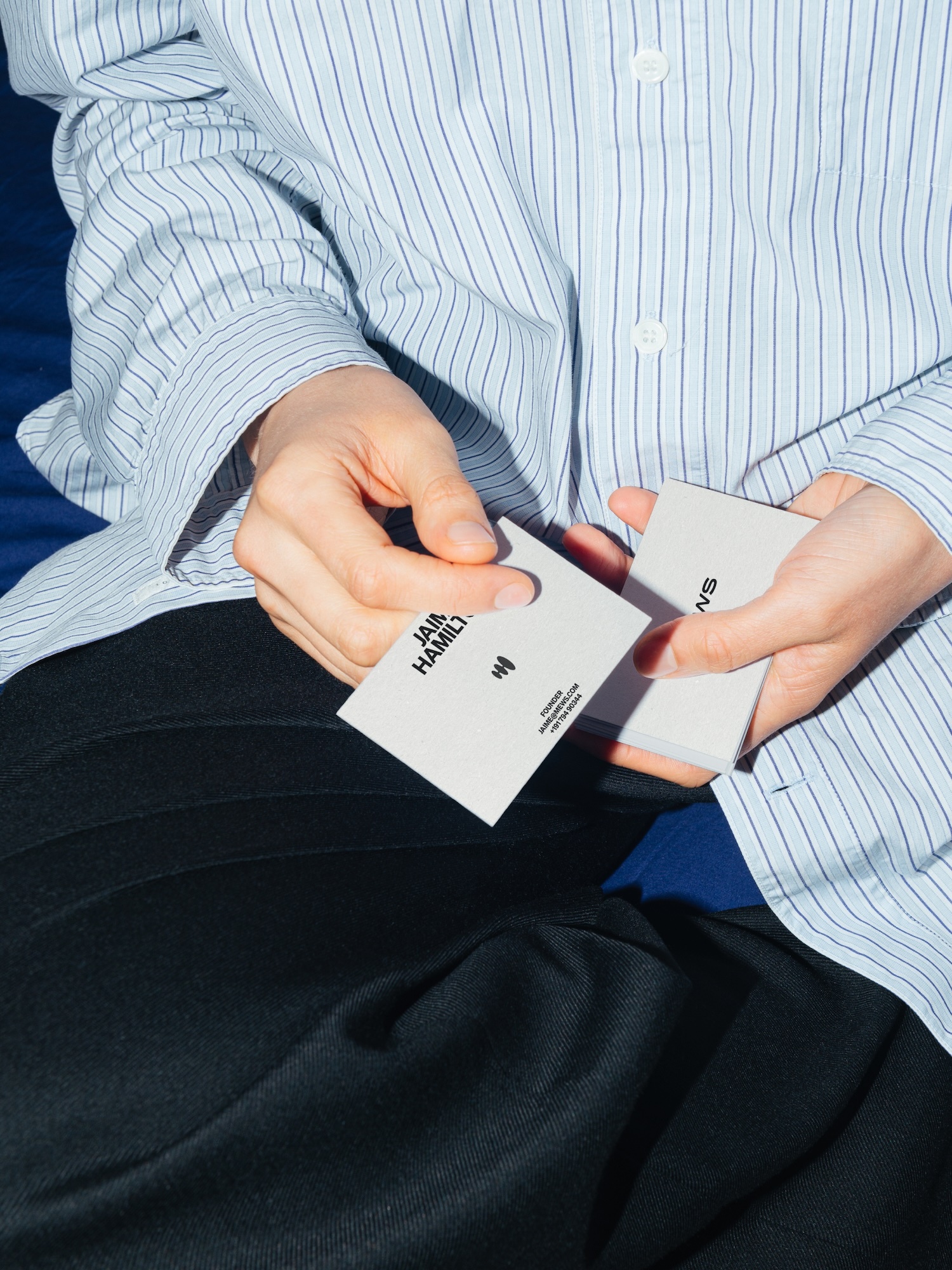 Close-up of adult hands holding two business cards in front of a striped shirt and dark pants on a blue background.