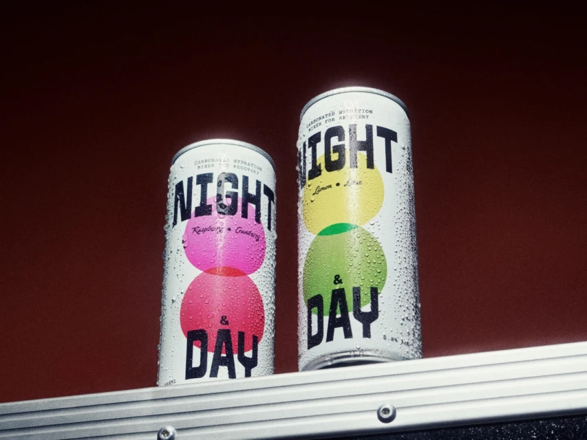 Two white aluminum cans of Night & Day brand carbonated hydration mixer on a metallic surface against a reddish-brown background.