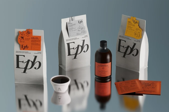 Three Ephemera Coffee bags, a bottle of cold brew coffee, a cup, and packaging on a reflective surface against a muted blue background.