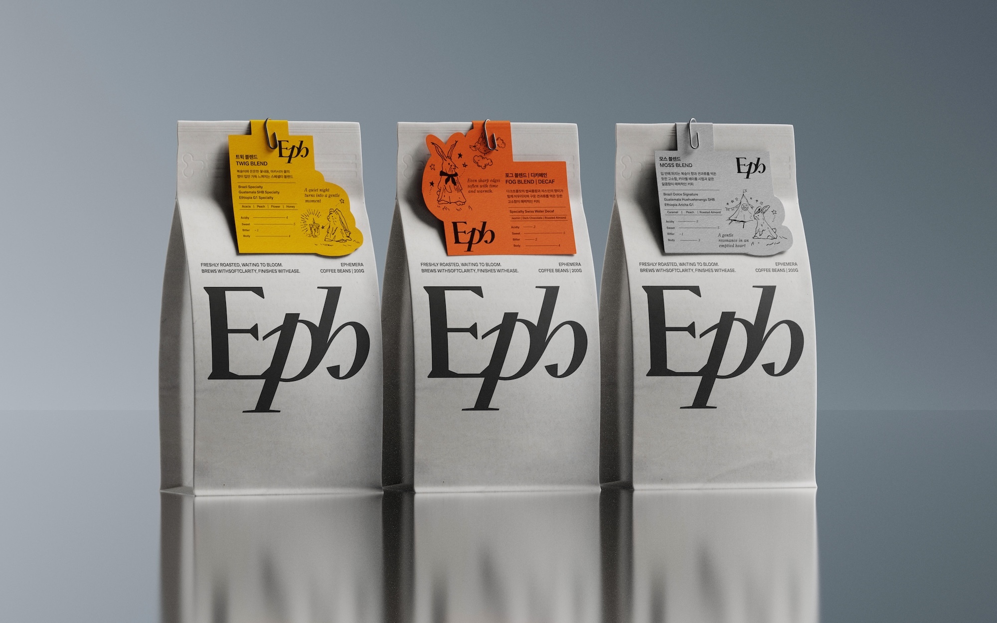 Three white bags of Ephermera coffee beans are displayed against a gray backdrop. Each has a uniquely colored tag attached.