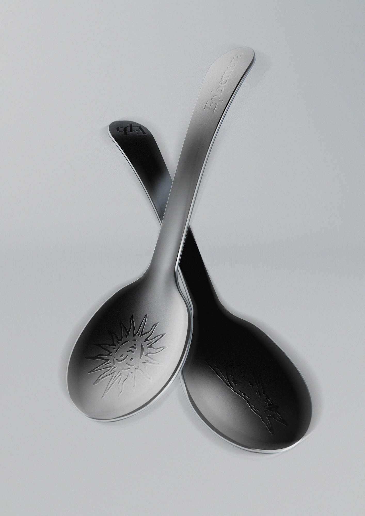 Two metal spoons, one with sun engraving, are crossed on a neutral surface. The spoons feature smooth, curved designs and reflective surfaces.