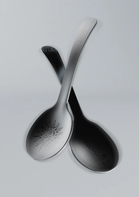 Two metal spoons, one with sun engraving, are crossed on a neutral surface. The spoons feature smooth, curved designs and reflective surfaces.