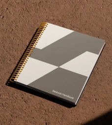 A spiral-bound notebook with a geometric grey and white cover resting on a textured brown earth surface.